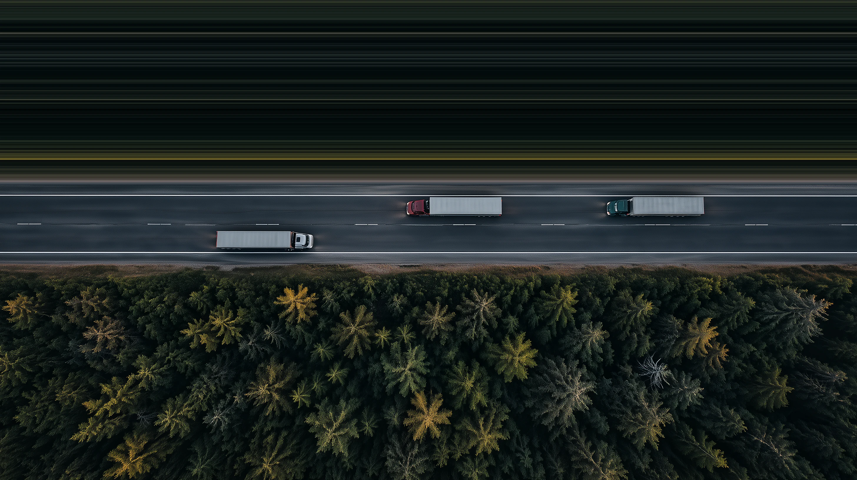 Aerial view of three semi trucks on a highway beside a pine forest