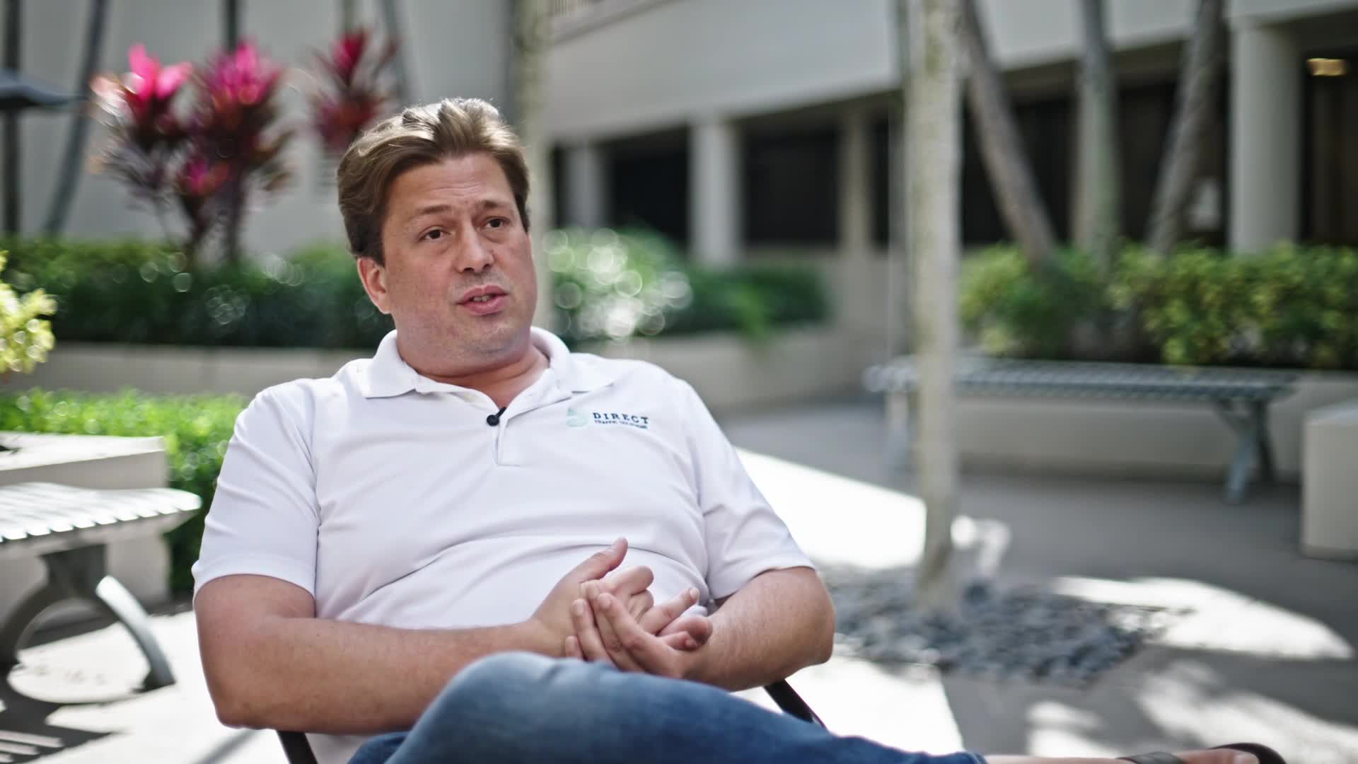 Christopher Griffin of Direct Traffic Solutions speaking on camera, seated in a sunlit courtyard